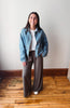 A woman stands in a room with a white wall and wooden floor, wearing a light blue denim jacket over a white t-shirt and brown wide-leg pants.