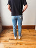A person viewed from behind wearing light blue slim-fit jeans, a black t-shirt, and white sneakers, standing on a wooden floor against a white wall.
