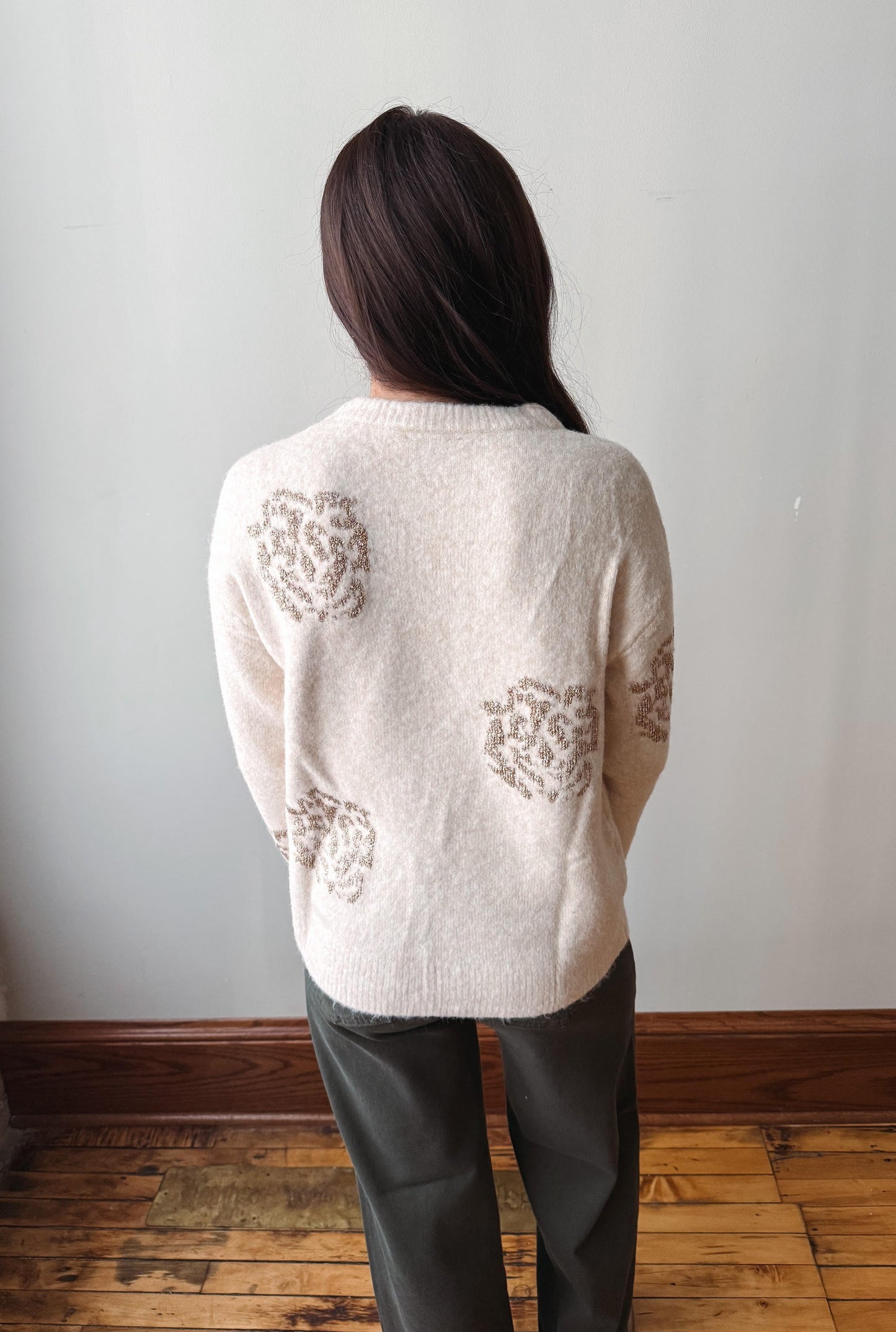 A woman viewed from behind wearing a cream-colored fuzzy knit sweater with brown textured floral patterns on the back, paired with dark gray pants, standing in a room with a light gray wall and wooden floor.