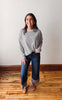 Woman wearing a gray striped sweater and blue jeans standing on a wooden floor.