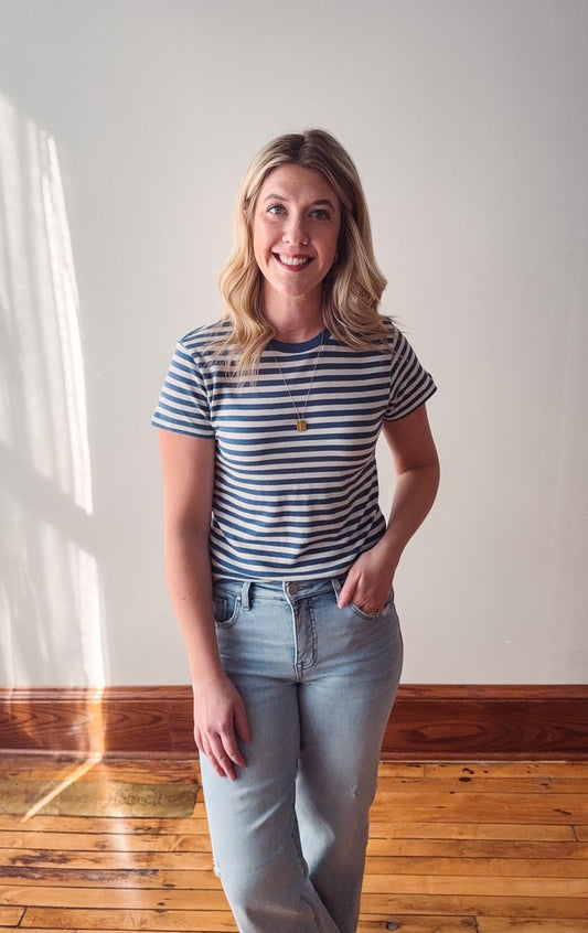 Woman wearing a striped shirt and jeans standing in a room with wooden flooring and a white wall.