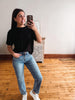 A woman taking a mirror selfie in a room with hardwood floors and a white wall, wearing a black short-sleeve t-shirt, light wash blue jeans, and white sneakers.