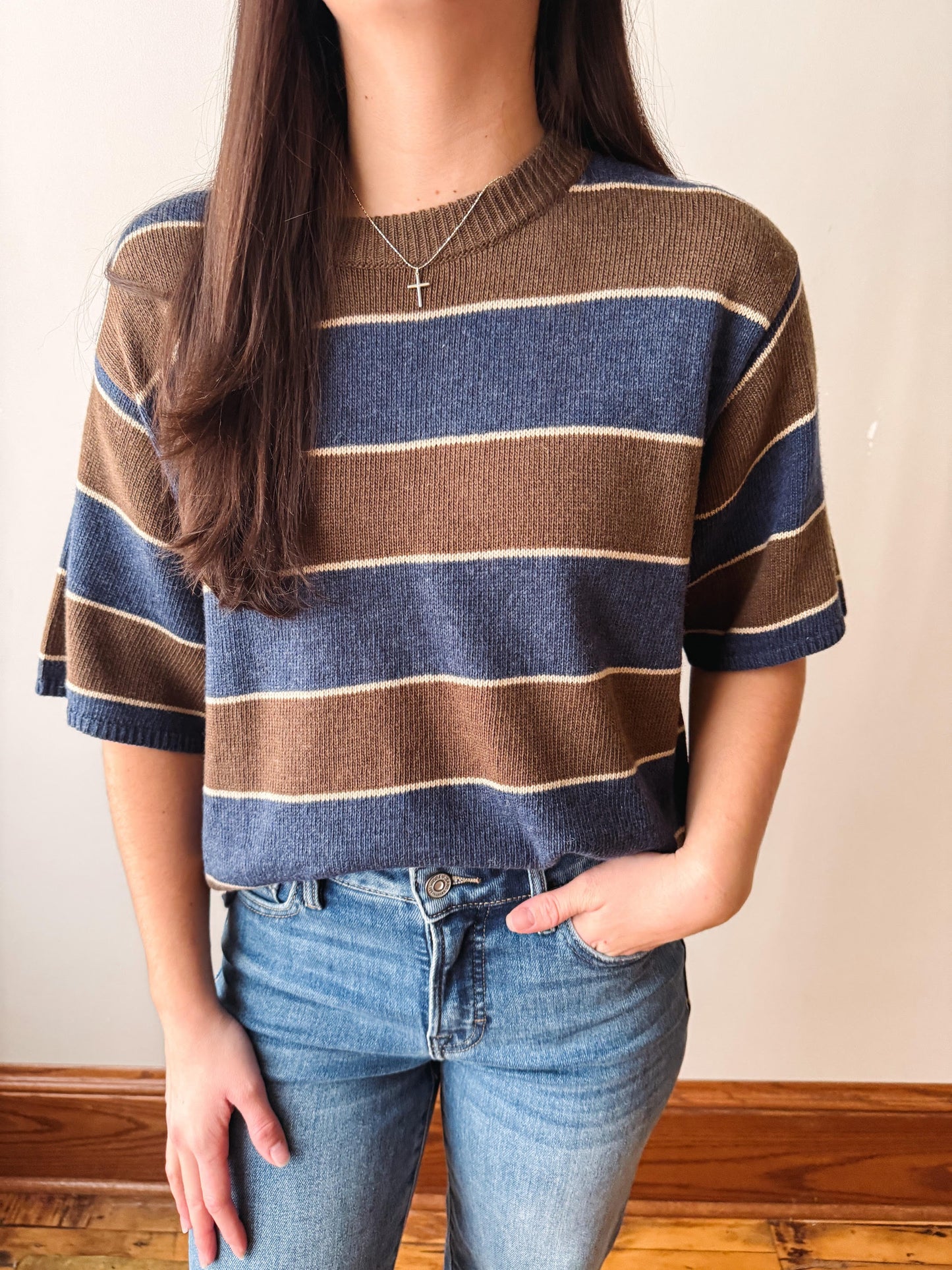 A person wearing a short-sleeve knit sweater with horizontal brown, navy blue, and cream stripes, paired with blue jeans, standing indoors against a plain light wall.