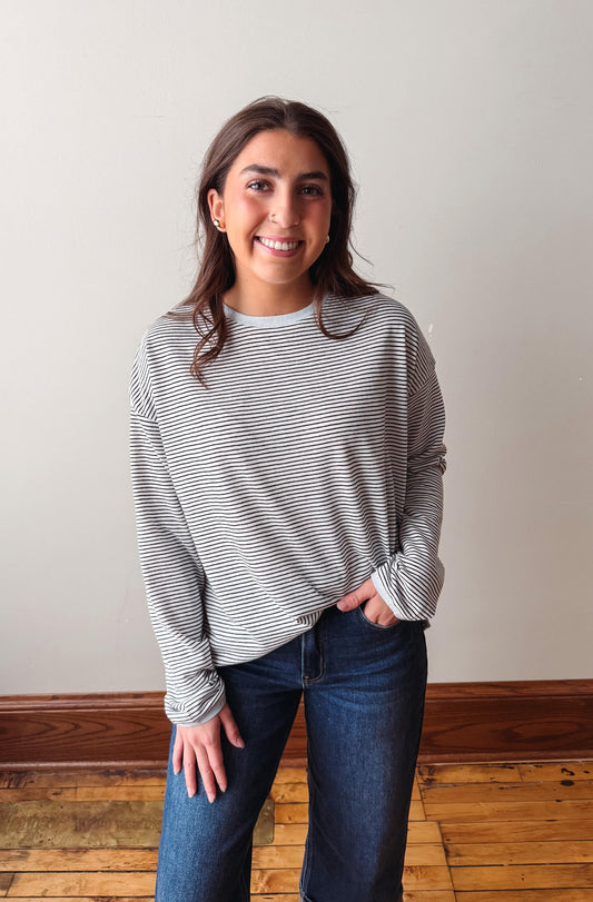 Woman wearing a striped long-sleeve shirt and jeans standing on a wooden floor with a white wall background.