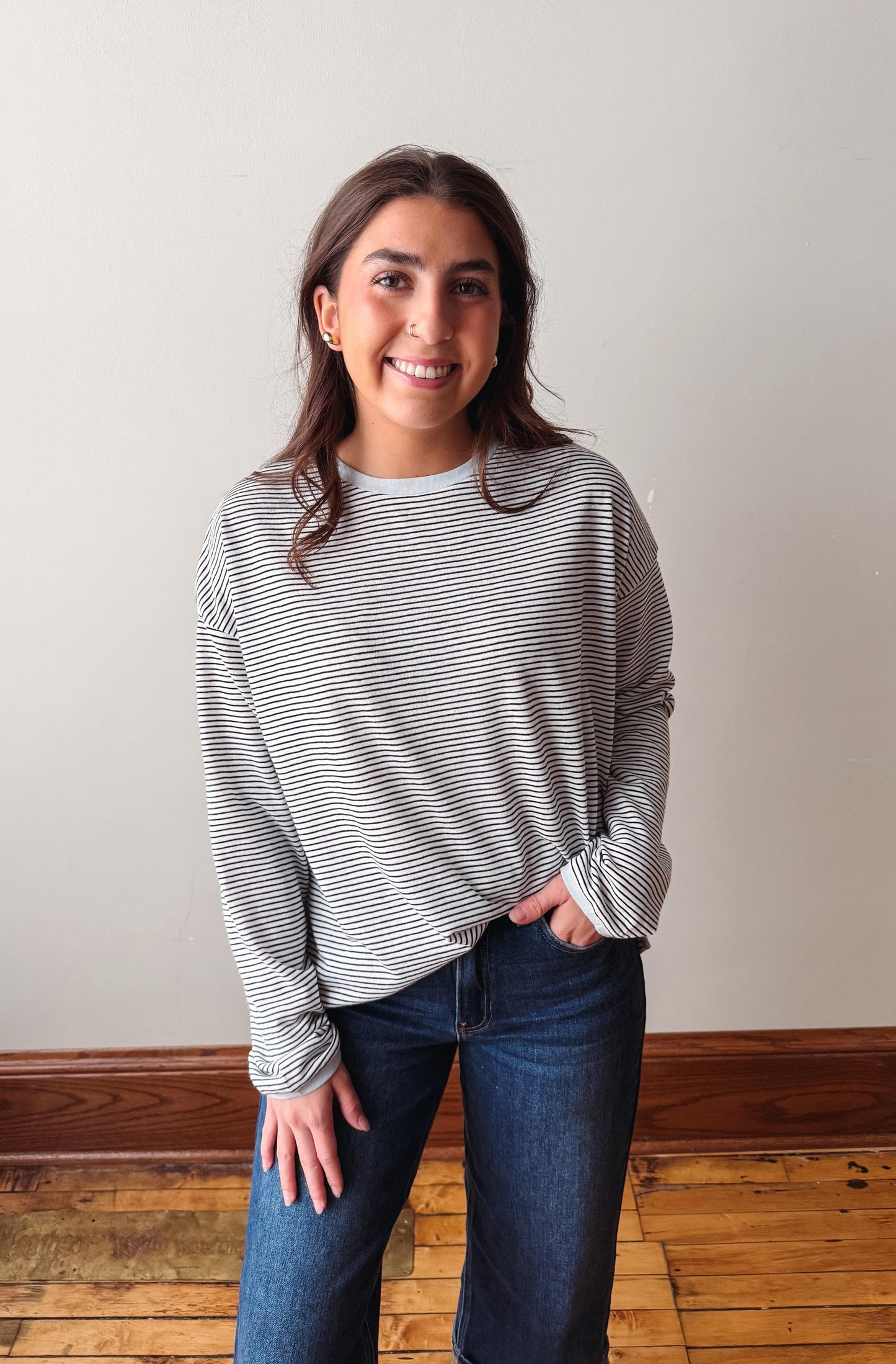 Woman wearing a striped long-sleeve shirt and jeans standing on a wooden floor with a white wall background.