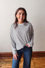 Woman wearing a striped long-sleeve shirt and jeans standing on a wooden floor with a white wall background.