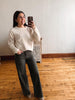 A woman takes a mirror selfie in a room with hardwood floors and a white wall, wearing a cream knit sweater with brown floral patterns and dark charcoal wide-leg pants.