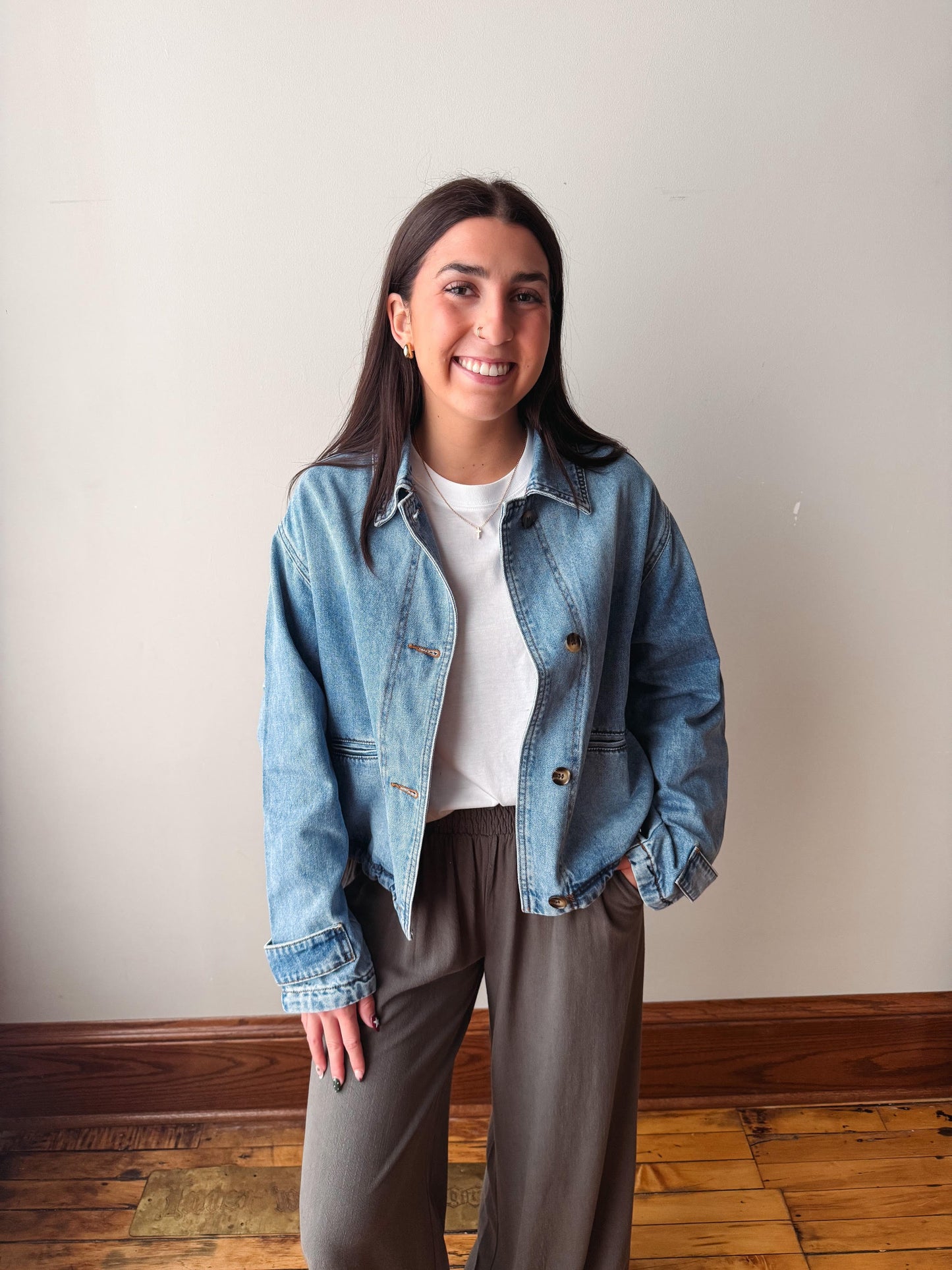 A woman smiling and posing indoors wearing a light blue cropped denim jacket over a white t-shirt and brown wide-leg pants, standing against a plain off-white wall with a wooden floor visible.