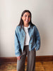 A woman smiling and posing indoors wearing a light blue cropped denim jacket over a white t-shirt and brown wide-leg pants, standing against a plain off-white wall with a wooden floor visible.