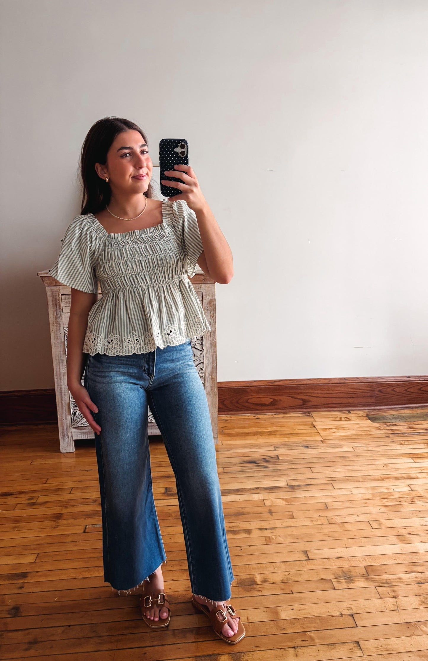 Woman taking a mirror selfie wearing a light green top and blue jeans on a wooden floor.