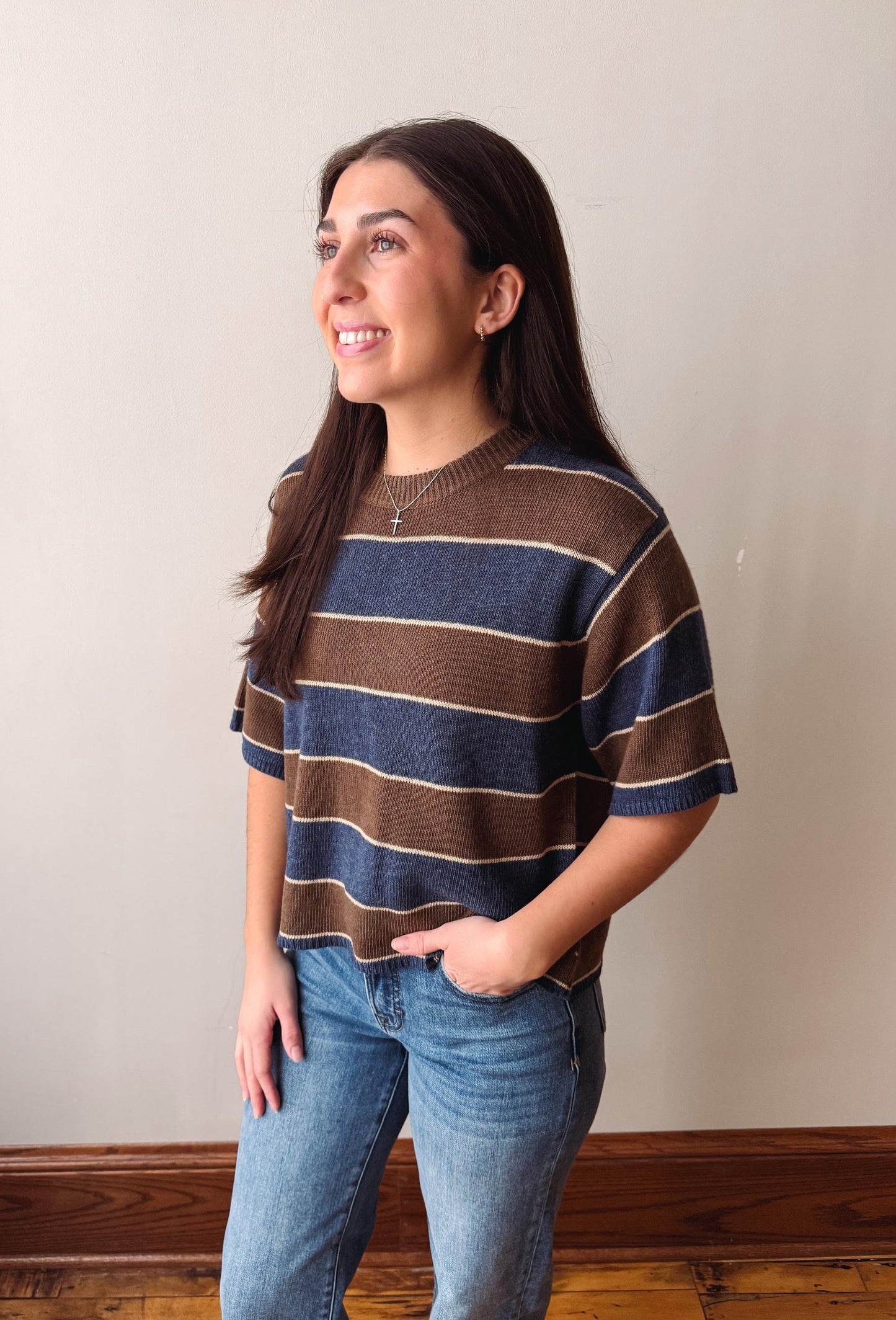 A woman wearing a brown and navy blue striped knit sweater with beige stripes, paired with blue jeans, standing against a plain beige wall.