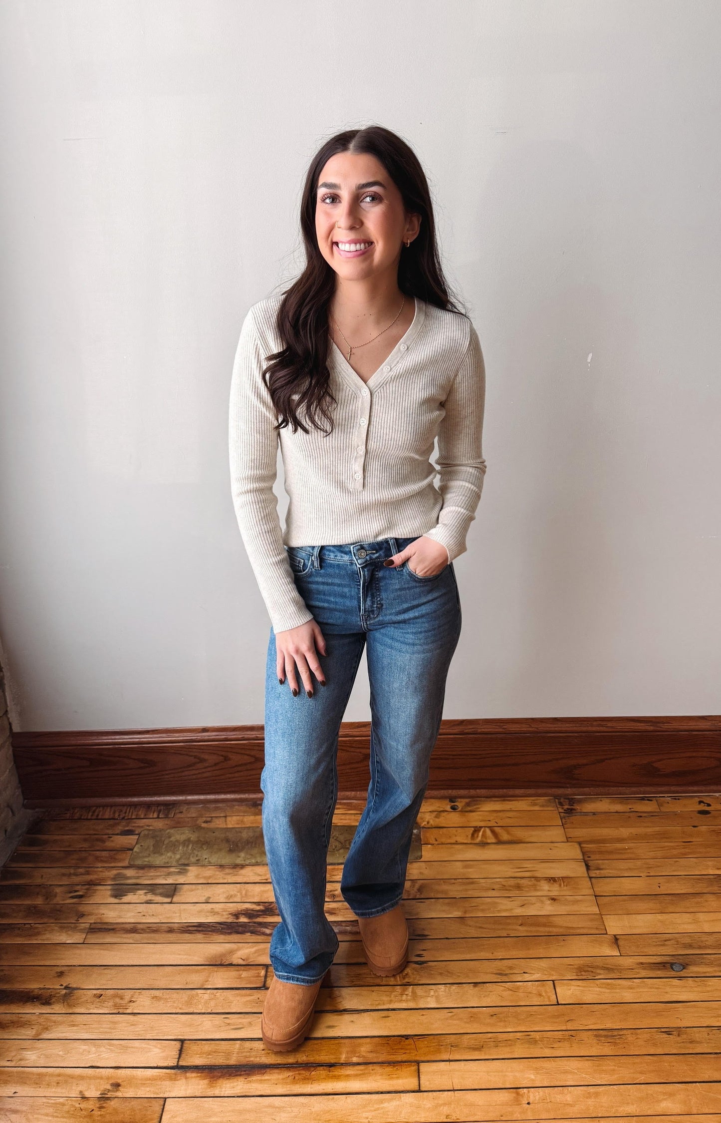 A woman stands in a room with a white wall and wooden floor, wearing a light beige knit cardigan, blue jeans, and tan boots.
