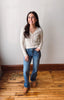 A woman stands in a room with a white wall and wooden floor, wearing a light beige knit cardigan, blue jeans, and tan boots.