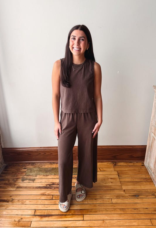 A woman modeling a brown sleeveless top and matching wide-leg pants set, standing on a wooden floor against a plain white wall.
