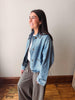 A woman wearing a light blue cropped denim jacket over a white t-shirt and gray wide-leg pants, standing in a room with a beige wall and wooden floor.