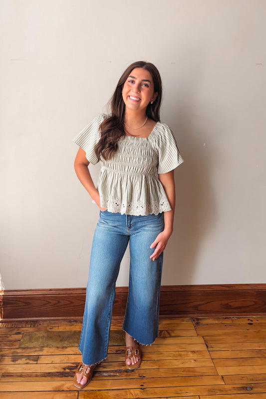 Woman wearing a light-colored top and blue jeans standing on a wooden floor against a plain wall.