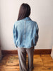 A woman viewed from behind wearing a light blue cropped denim jacket and brown wide-leg trousers, standing in a room with a light-colored wall and wooden floor.