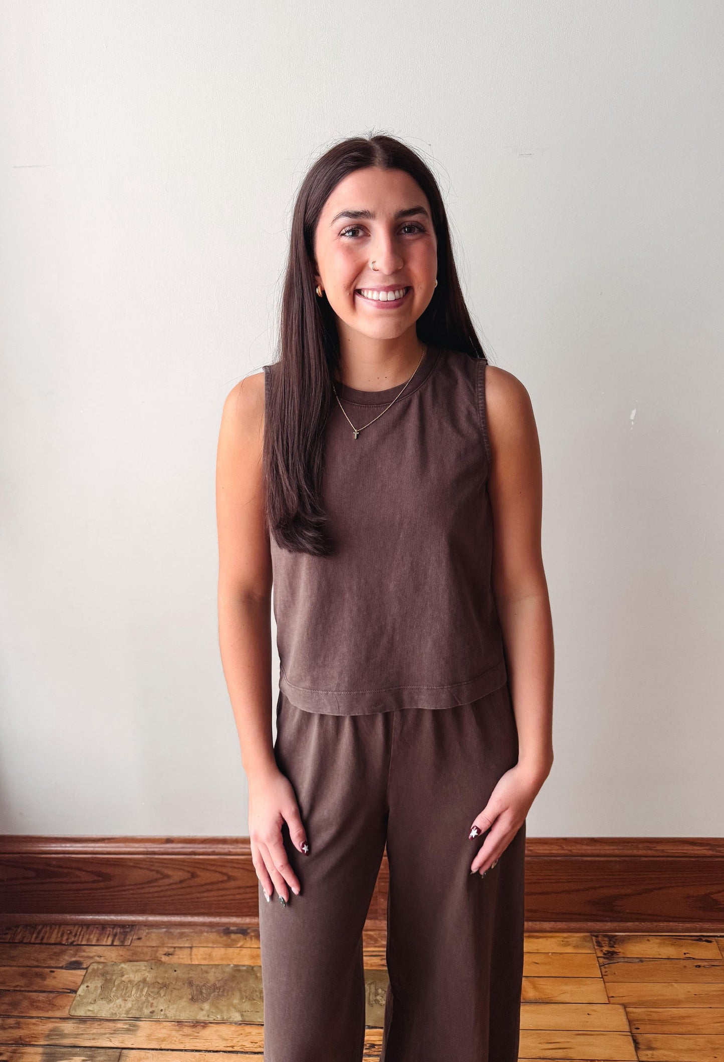 A woman with long dark hair stands indoors wearing a matching sleeveless cropped top and wide-leg pants set in a muted brown color. She is positioned in front of a plain off-white wall with a wooden baseboard and wood floorboards visible.