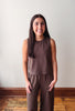 A woman with long dark hair stands indoors wearing a matching sleeveless cropped top and wide-leg pants set in a muted brown color. She is positioned in front of a plain off-white wall with a wooden baseboard and wood floorboards visible.