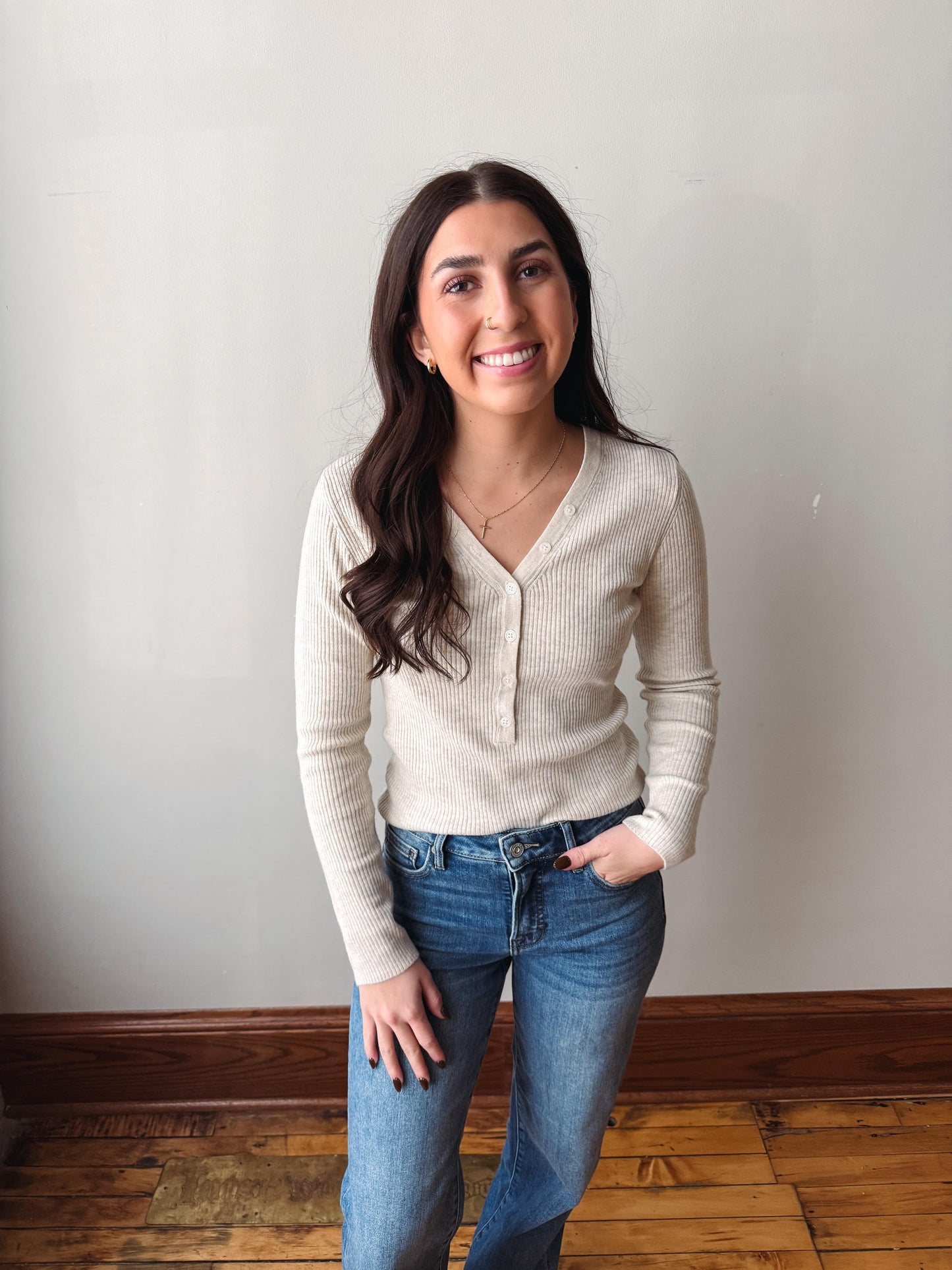A smiling young woman with long dark hair stands indoors wearing a ribbed cream-colored long-sleeve henley top and blue jeans against a plain light gray wall and wooden floor.