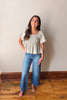 Woman wearing a light-colored top and blue jeans standing on a wooden floor against a plain wall.