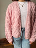 A person wearing a chunky pink cable-knit cardigan over a white t-shirt and light blue jeans, standing indoors against a neutral wall.