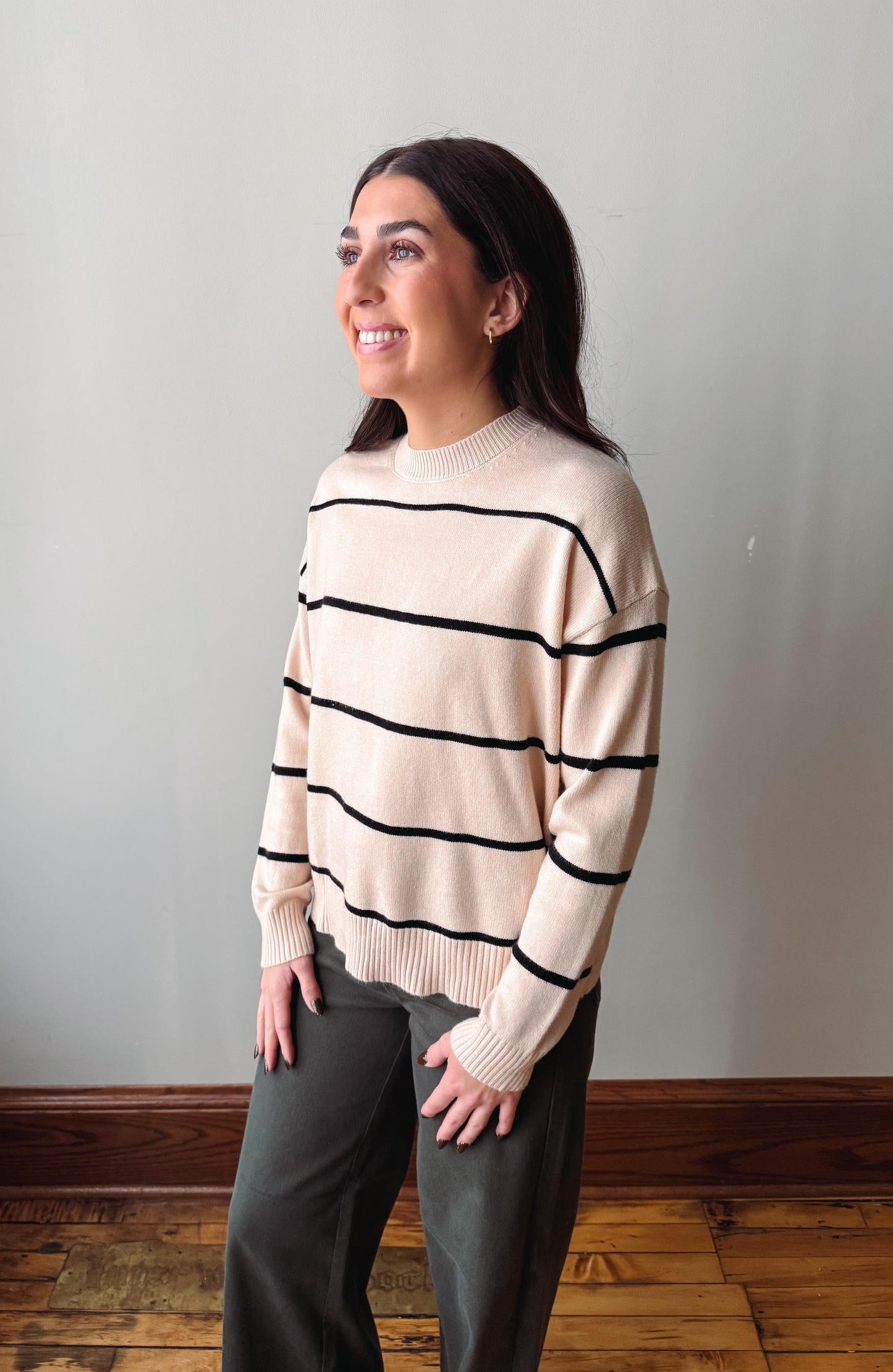 A woman wearing a cream-colored knit sweater with black horizontal stripes and dark gray pants, standing in a room with a light gray wall and wooden floor.