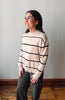 A woman wearing a cream-colored knit sweater with black horizontal stripes and dark gray pants, standing in a room with a light gray wall and wooden floor.