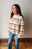 A woman wearing a fuzzy, long-sleeve knit sweater with horizontal stripes in cream, tan, and dark brown, paired with blue jeans, standing against a neutral beige wall and wooden floor.