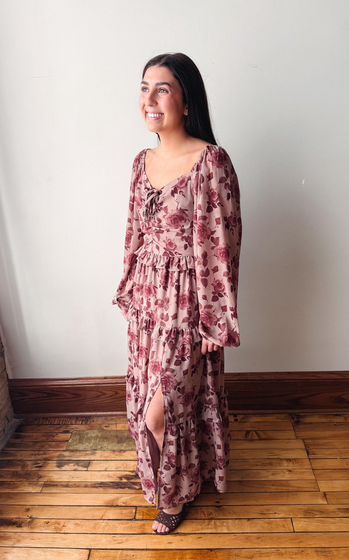 A woman wearing a long-sleeved, dusty rose floral maxi dress with a tie-neck detail and side slit, standing on a wooden floor against a plain white wall.