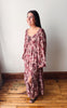 A woman wearing a long-sleeved, dusty rose floral maxi dress with a tie-neck detail and side slit, standing on a wooden floor against a plain white wall.