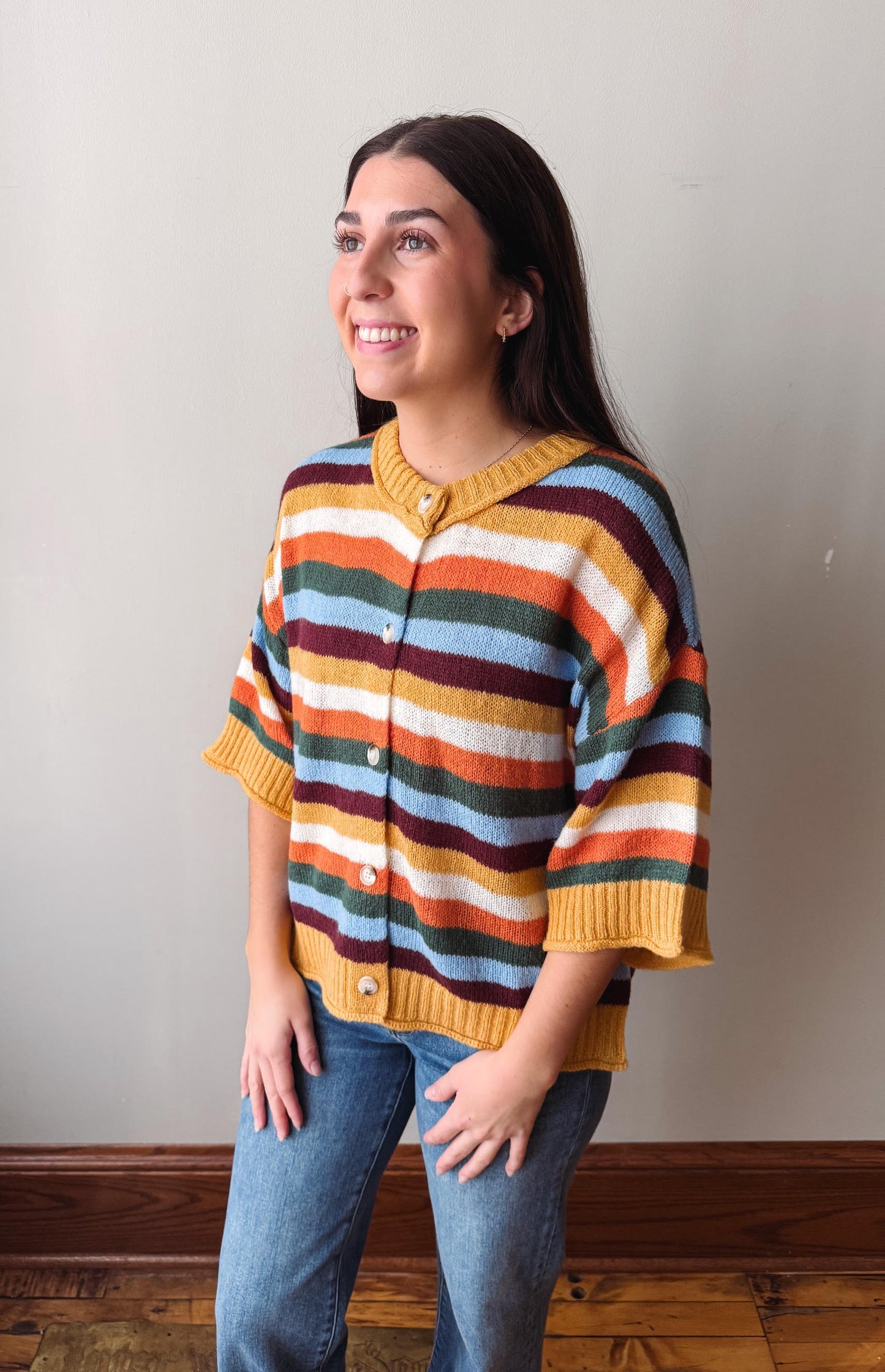 A woman wearing a colorful striped knit cardigan sweater with blue jeans, standing against a plain light gray wall.