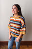 A woman wearing a colorful striped knit cardigan sweater with blue jeans, standing against a plain light gray wall.