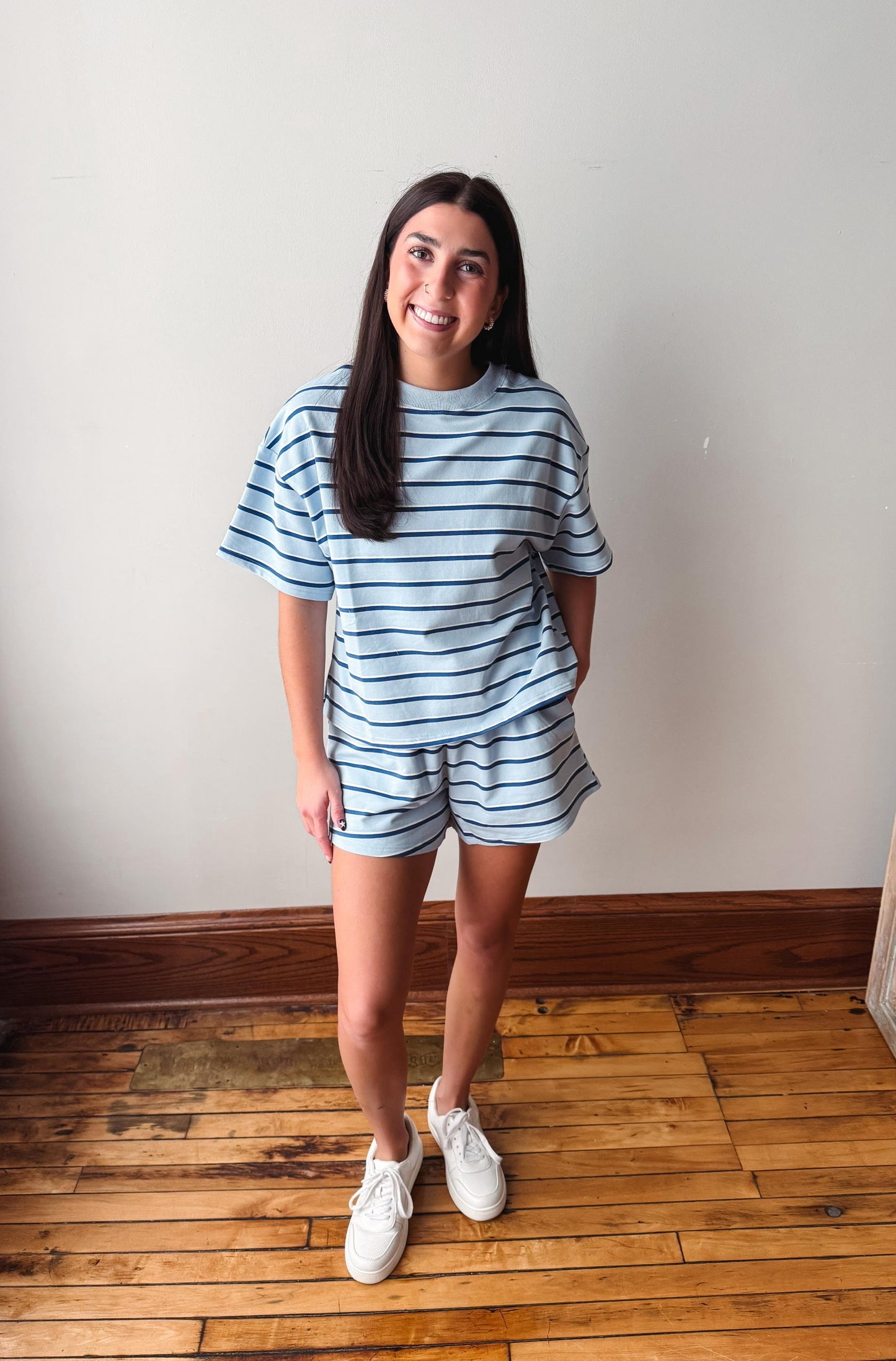 A woman modeling a matching light blue and navy striped lounge set, consisting of a short-sleeve t-shirt and shorts, paired with white sneakers, standing in a room with a white wall and wooden floor.