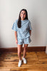 A woman modeling a matching light blue and navy striped lounge set, consisting of a short-sleeve t-shirt and shorts, paired with white sneakers, standing in a room with a white wall and wooden floor.