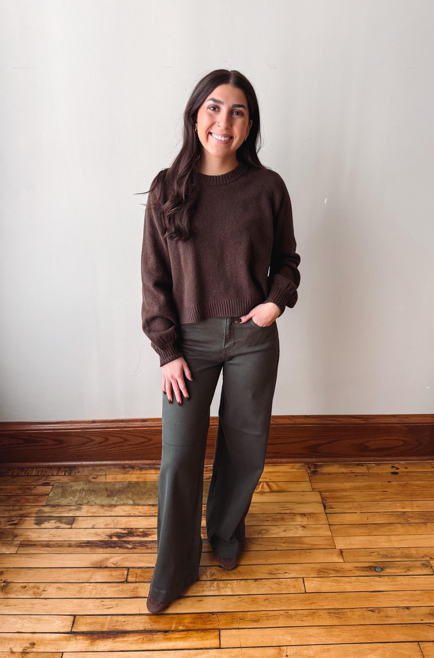 A woman stands in a room with a light gray wall and wooden floor, wearing a dark brown long-sleeve knit sweater and dark gray wide-leg pants.