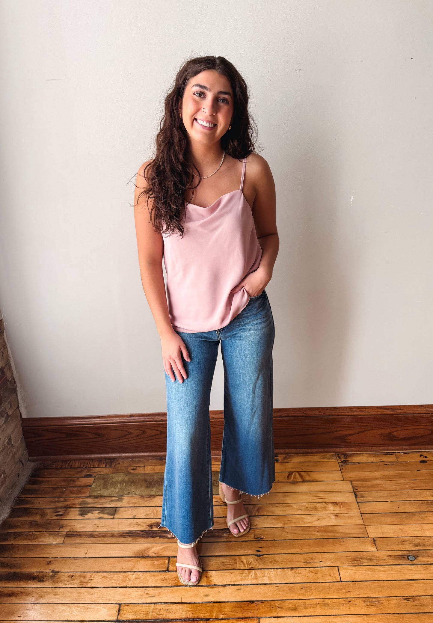 Woman wearing a pink top and blue jeans standing on a wooden floor against a white wall.