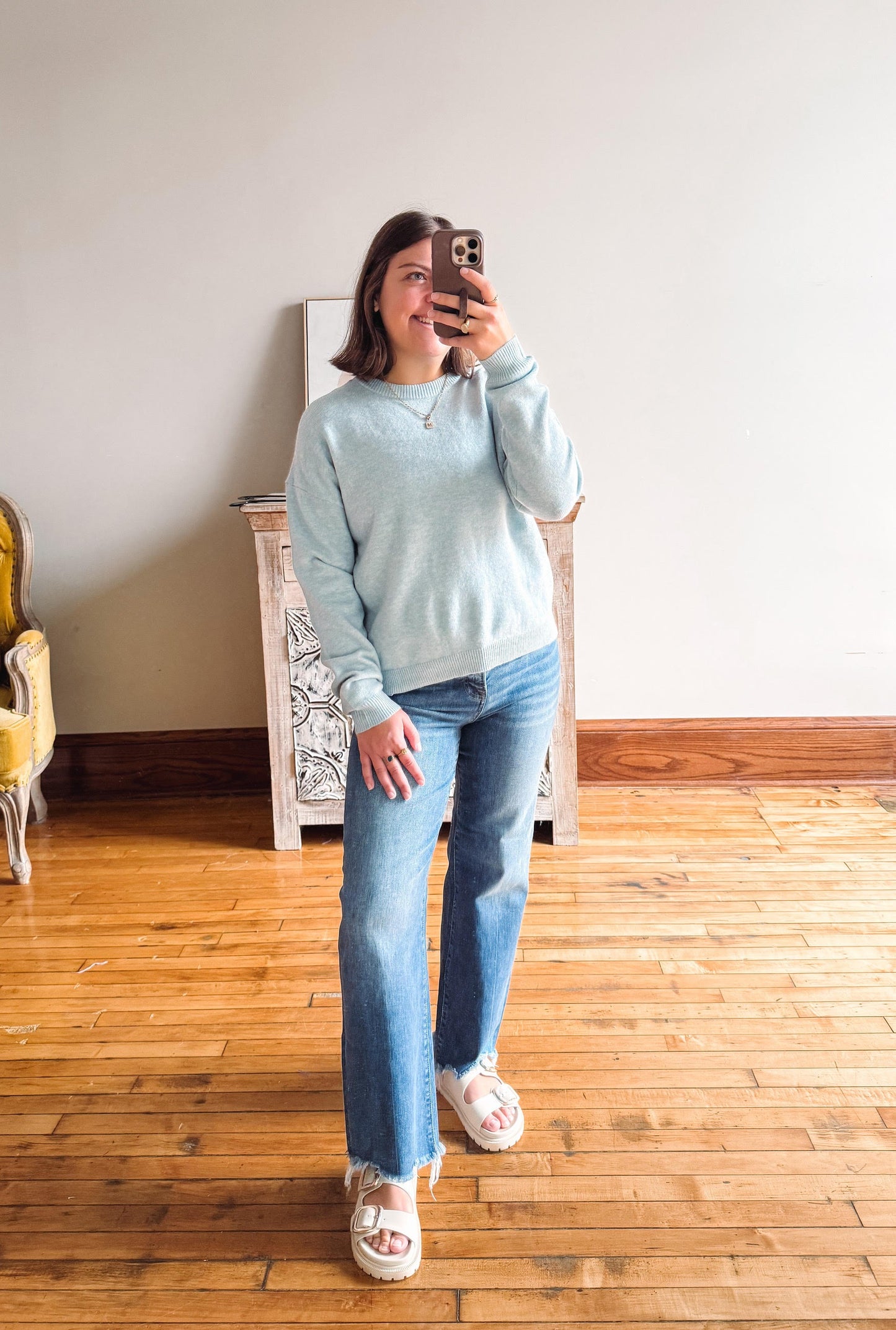 A woman takes a mirror selfie wearing a light blue long-sleeve crewneck sweater, blue flare jeans, and white slide sandals in a room with wooden floors and a white wall.