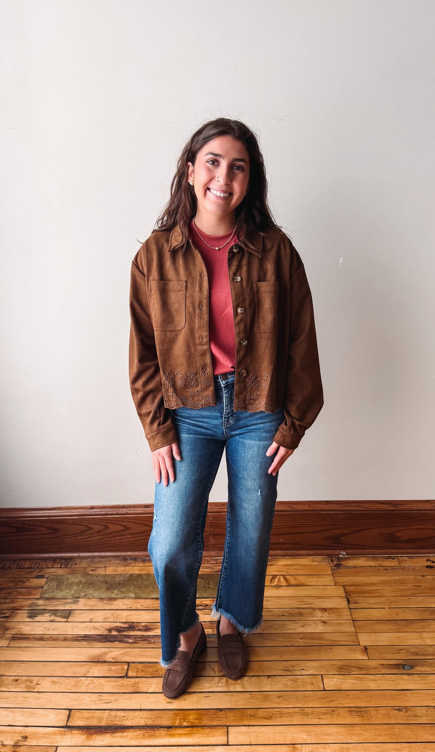 A woman stands in a room with a white wall and wooden floor, wearing a brown suede jacket over a dusty rose top, blue jeans, and brown suede loafers.