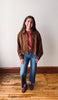 A woman stands in a room with a white wall and wooden floor, wearing a brown suede jacket over a dusty rose top, blue jeans, and brown suede loafers.