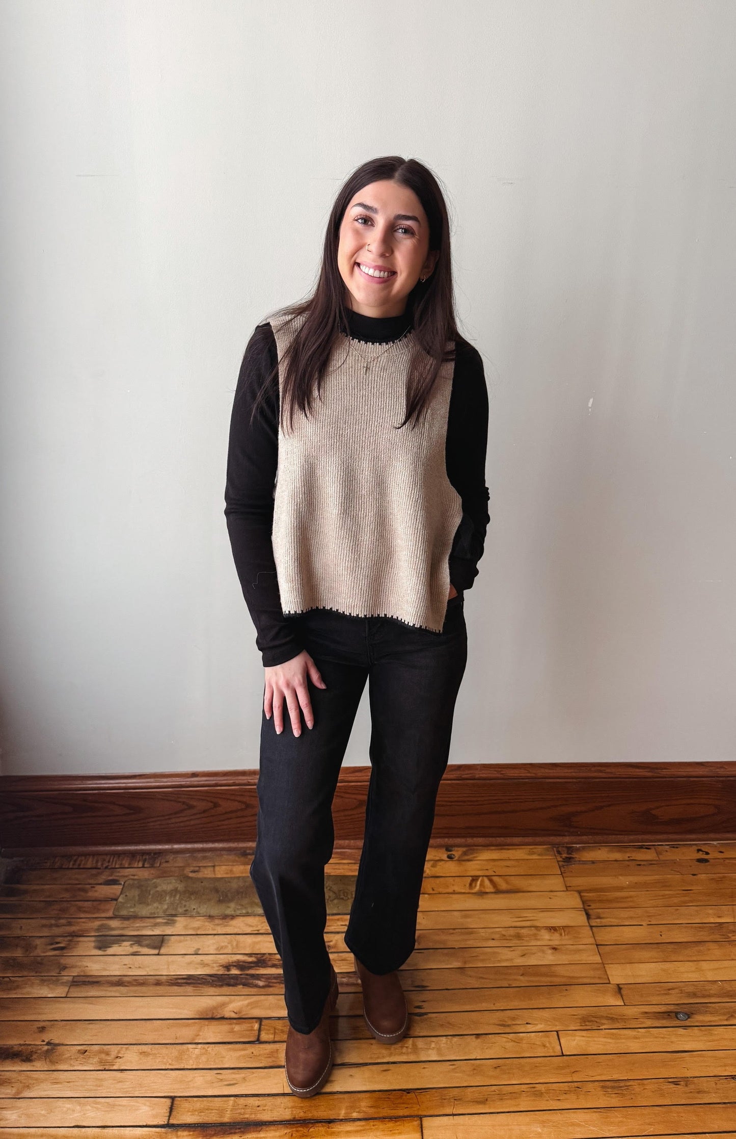 A woman modeling a sleeveless beige knit sweater vest over a black long-sleeve mock neck top, paired with black pants and brown boots, standing on a wooden floor against a plain off-white wall.