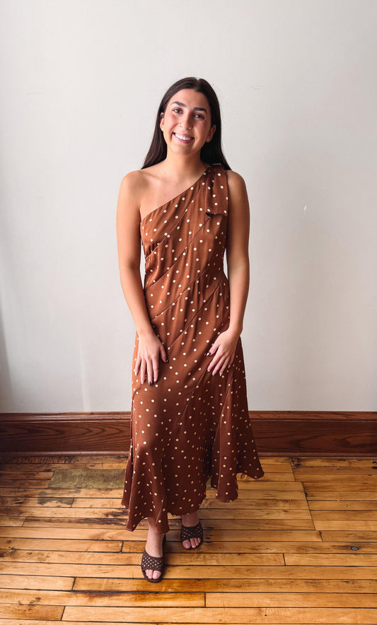 A woman wearing a brown one-shoulder maxi dress with white polka dots, standing on a wooden floor against a plain off-white wall.