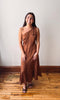 A woman wearing a brown one-shoulder maxi dress with white polka dots, standing on a wooden floor against a plain off-white wall.