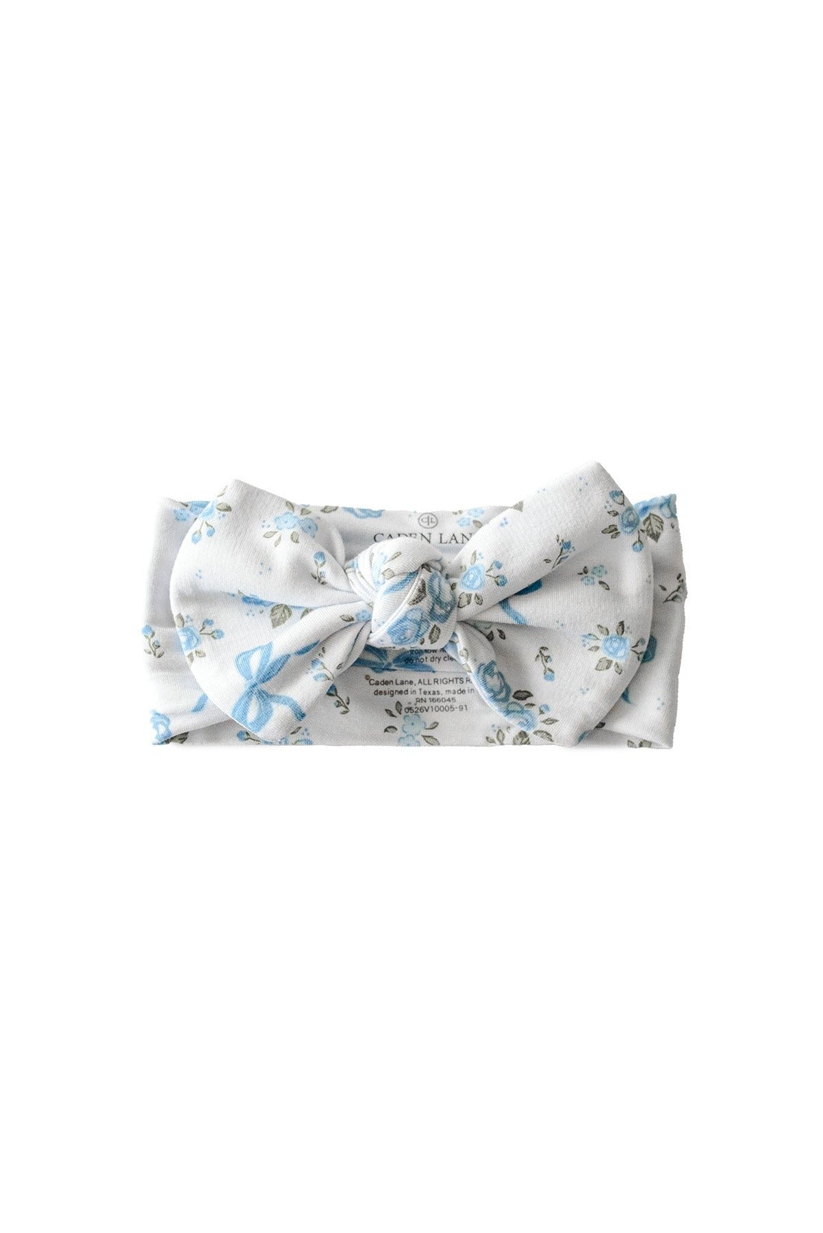 A white fabric headband with a large bow, featuring a delicate light blue floral pattern and a printed brand label in the center, shown on a plain white background.