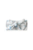 A white fabric headband with a large bow, featuring a delicate light blue floral pattern and a printed brand label in the center, shown on a plain white background.