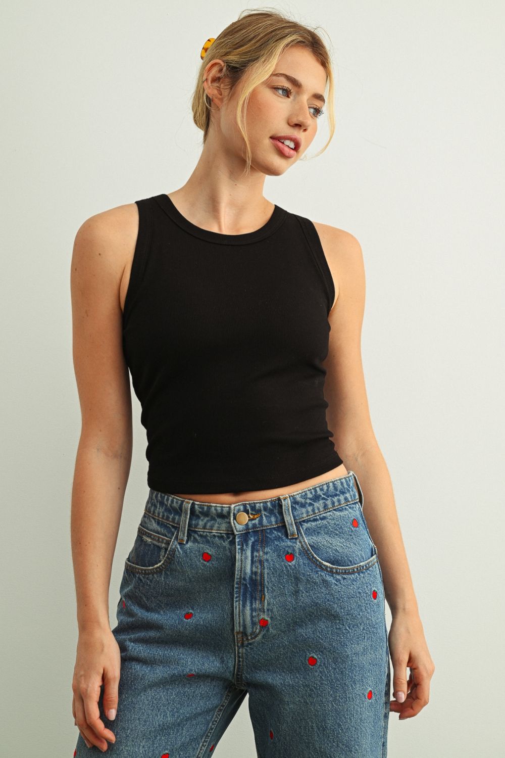 Woman wearing a black sleeveless top and blue jeans with red buttons on a plain background