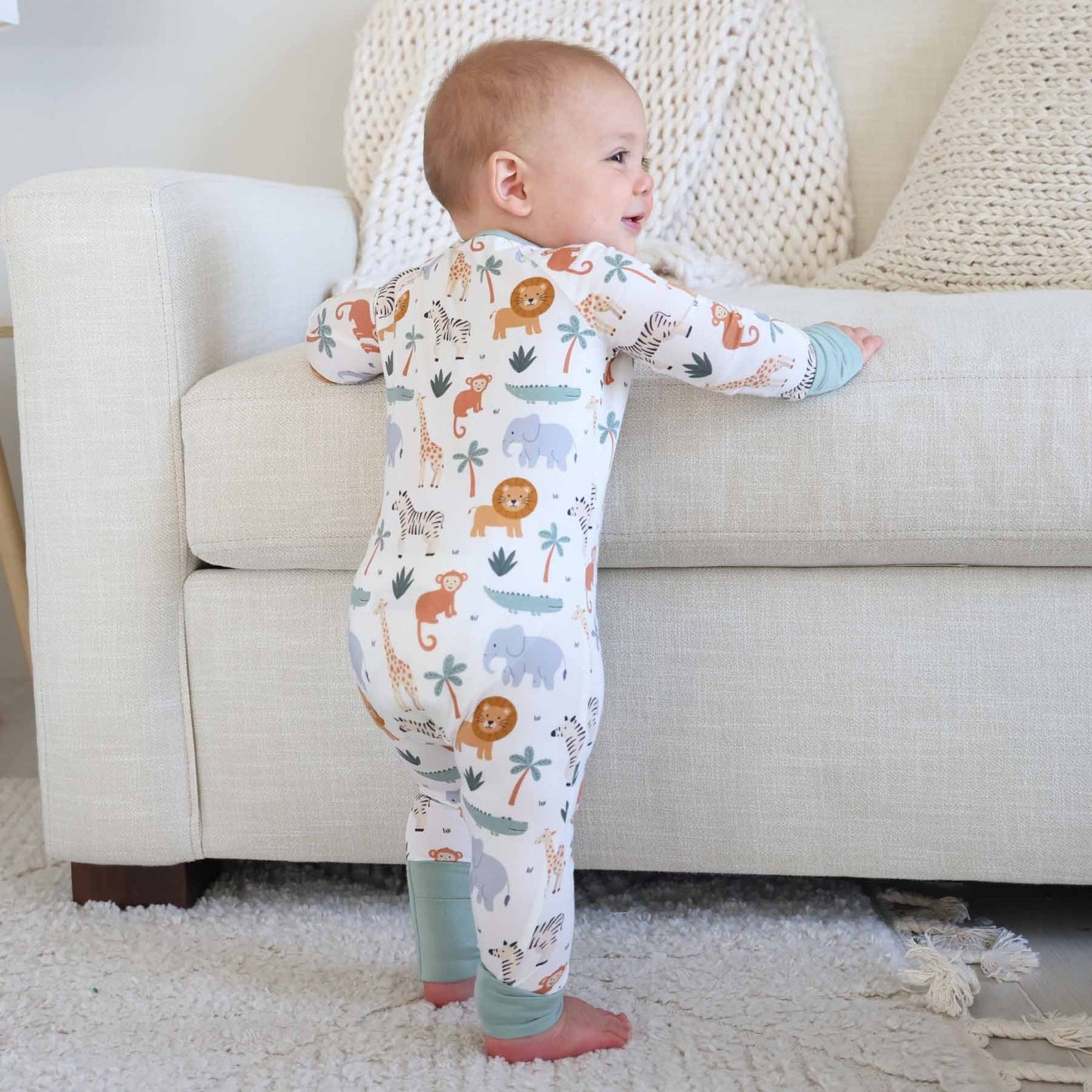 Baby in a onesie with animal prints standing in a living room.