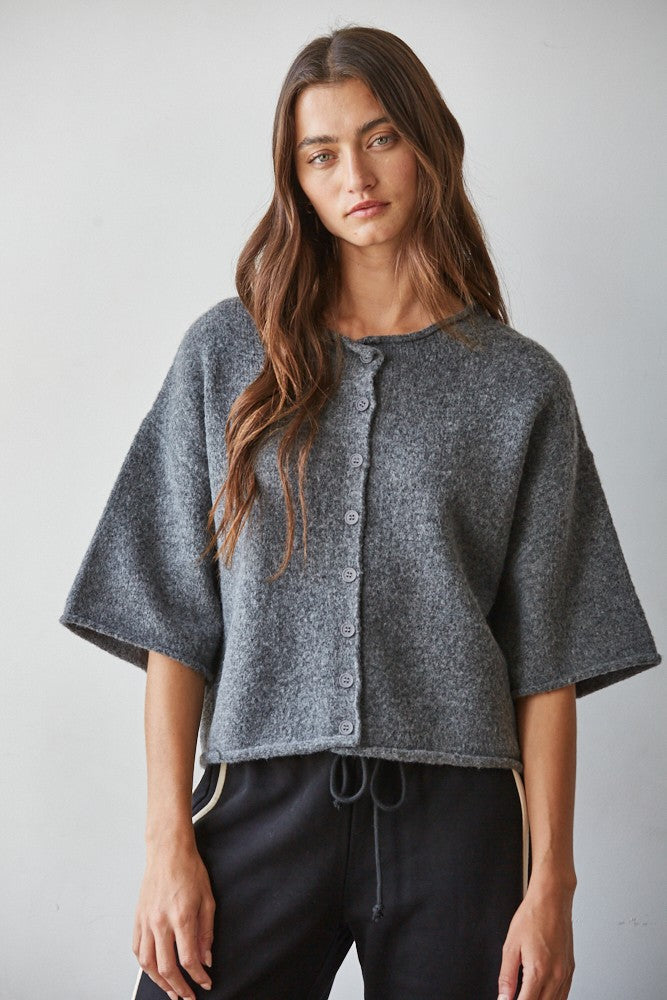 A woman wearing a cropped, short-sleeved gray knit cardigan with a button front, photographed against a plain light gray studio background.