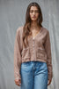 A woman modeling a cropped, dusty rose-brown fuzzy cardigan with white contrast stitching and button details, paired with high-waisted blue jeans, set against a textured gray backdrop.