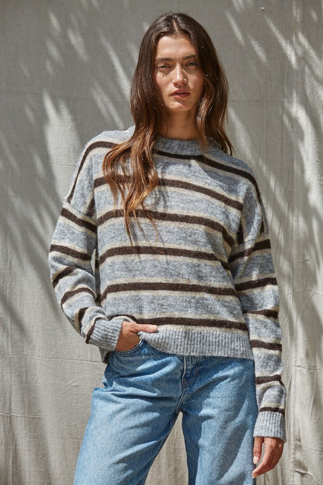 A woman wearing a gray and brown horizontally striped knit sweater with light blue jeans, standing against a textured beige wall with leaf shadows.